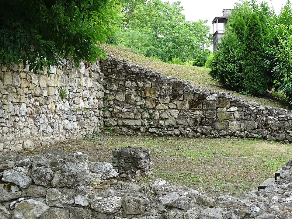 A wall of roughly squared stones about a person's height, stepping down along a grassy slope, with low wall stubs in the foreground and trees behind.