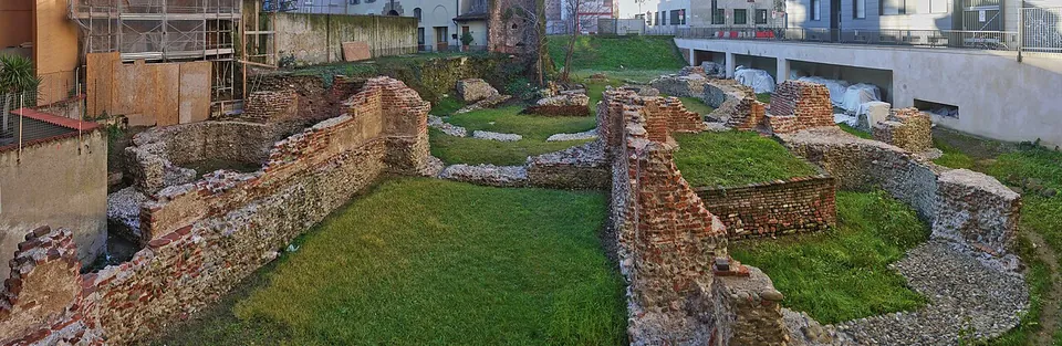 A panorama of brick-faced walls standing above the grass and dividing it into rooms, with a gravel path between them and the backs of town houses beyond.