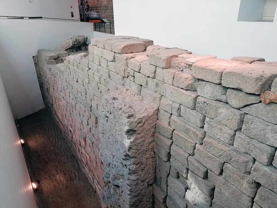 Seen from above, a massive wall of roughly squared grey tufa blocks laid in courses, stepped and broken at the top, rising from a narrow excavated trench beside a white museum wall.