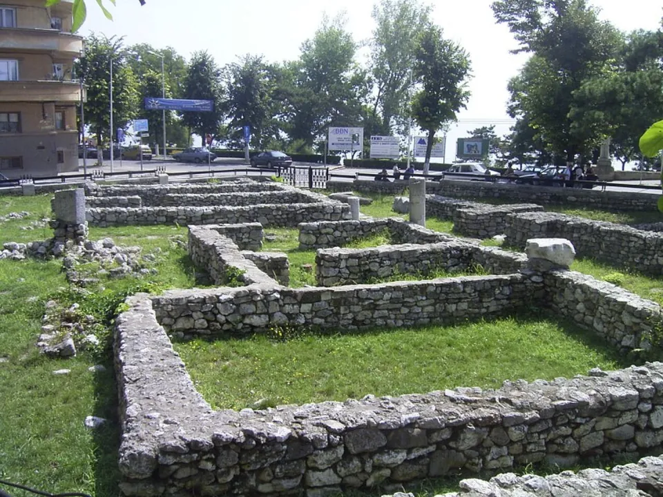 Low walls of rough stone outlining rooms in a mown lawn in the middle of a city, with a few stone fragments, trees, parked cars and a building behind.