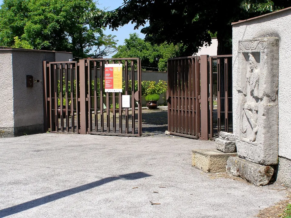 An open brown metal gate in a plastered wall with a red and yellow poster on it, a garden beyond, and a tall carved stone relief standing on a block beside the gate.
