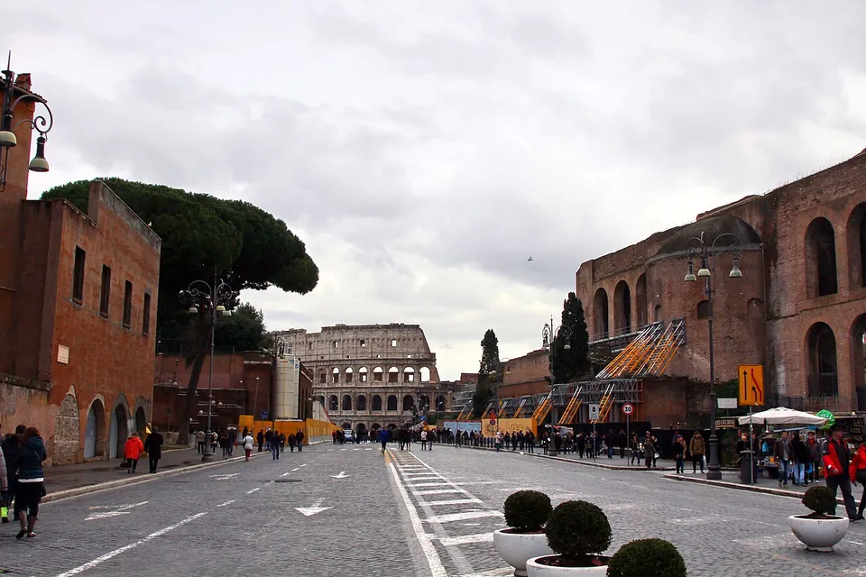A broad paved avenue with walkers and planters, brick buildings on either side, and the arches of the Colosseum closing the view at the far end under a grey sky.