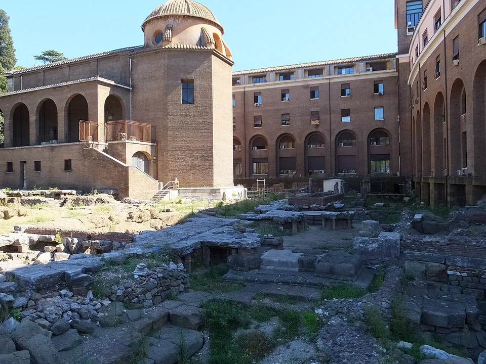 The whole excavation from one corner, courses of grey stone blocks and slabs at several levels, enclosed by a tall modern brick building with rows of arched windows and, at the far end, the church with its dome.