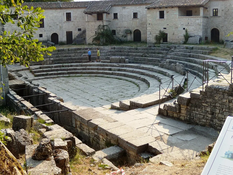 A semicircle of stone seating rows around a paved stone floor, with a curved range of two-storey stone farmhouses standing on top of the seating behind, and visitors on the steps.