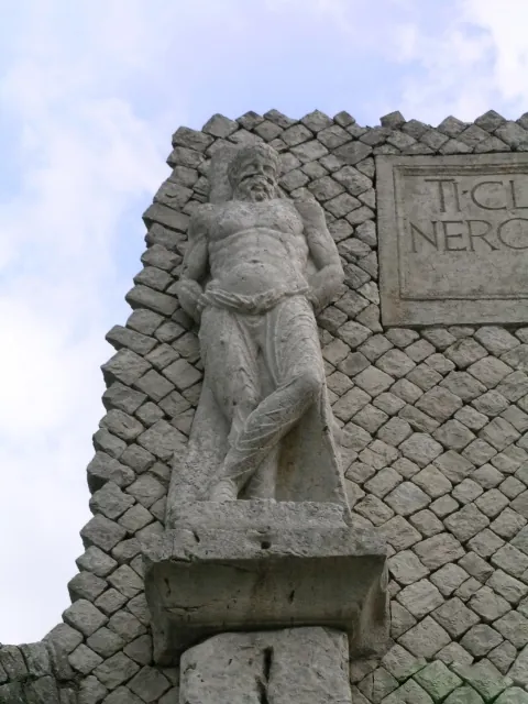 A weathered stone statue of a bearded, bare-chested man standing on a projecting bracket against a wall of small stones laid in a net pattern, beside a carved block with the letters TI CL and NERO.