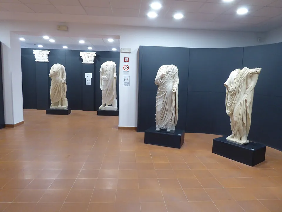 A museum room with a tiled floor and dark display walls, where four headless marble statues in draped robes stand on black plinths, with two carved capitals mounted on the wall behind.
