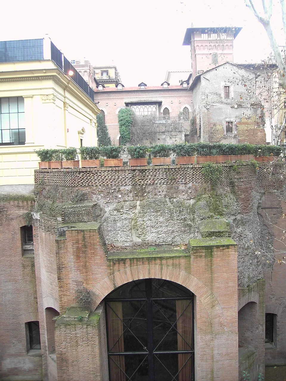 The Roman brick front of a hall, a large arched doorway closed by a glazed iron gate, with the rough concrete of a dome above it and a row of potted shrubs along the top; later buildings, one with a small tower, rise behind.