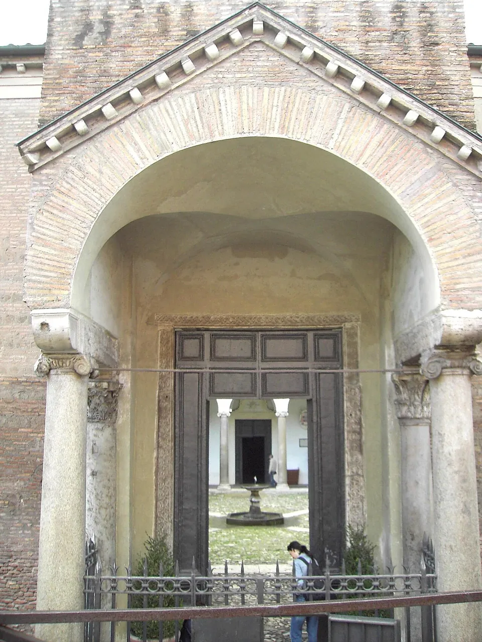 A brick gatehouse with a pediment over a deep round arch carried on two columns, an iron railing across the front, and through the open doors a sunlit courtyard with a fountain and the church facade beyond.