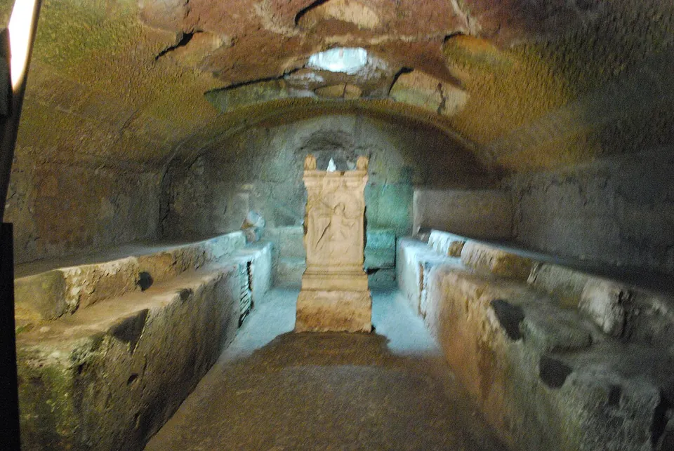 A low vaulted underground room with rough stone benches running along both side walls, and at the far end, lit pale against the dark, a carved stone altar standing on a base before a shallow apse; a small opening shows in the vault above.