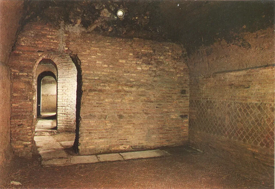 An underground room of red brick walls, a band of net-pattern masonry low on the right-hand wall, and a round-arched brick doorway on the left opening into a passage with further doorways beyond.