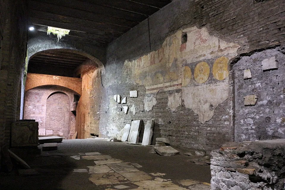 A long, low underground hall under a timber ceiling, its brick walls lit by lamps. On the right-hand wall patches of faded painted plaster survive, with a row of round yellow medallions; marble fragments lean against the foot of the wall, and at the far end a brick arch frames a bricked-up round-headed opening.