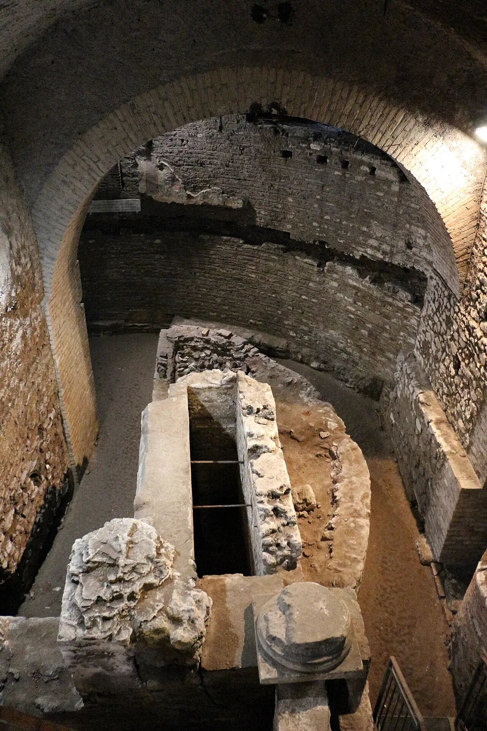 Looking down into a curved brick space under a vaulted modern ceiling. A narrow passage follows the inside of the curving wall, and in the middle stands a long, open-topped stone box, with a broken column base in the foreground.