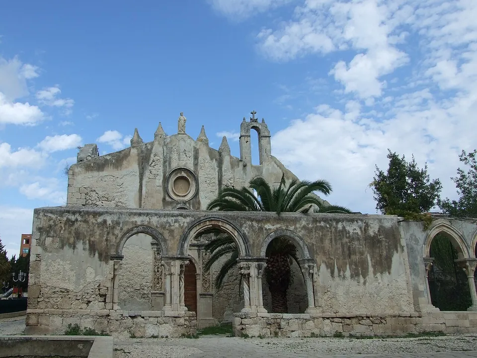 A roofless church of weathered limestone, with a gabled front pierced by a rose window and a small bell-cote on top, behind a lower wall of round arches on columns, with a palm tree in front.