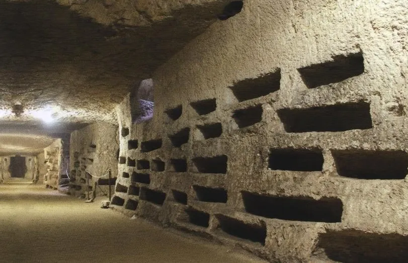 A rock wall cut with rows of small rectangular burial niches, stacked several high, along a lit gallery that runs into the distance.