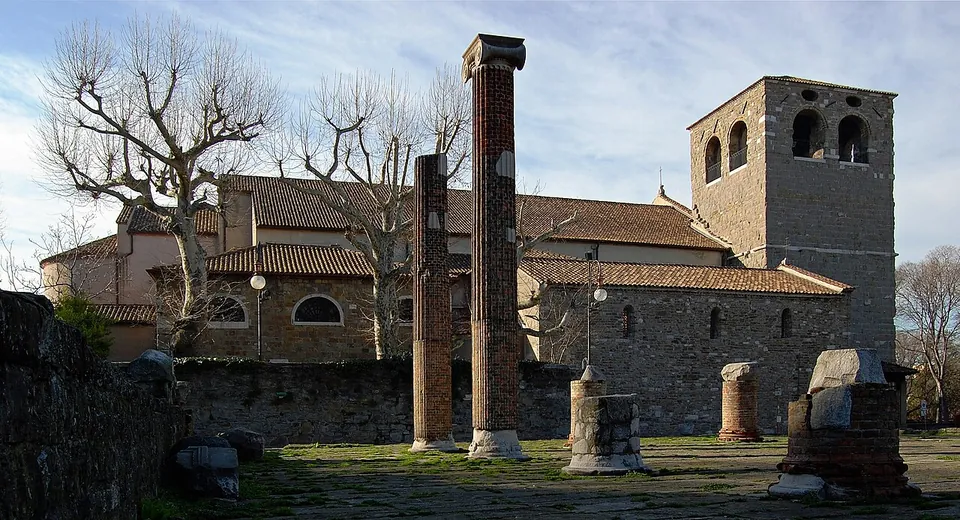 Two tall columns and a few stone blocks standing on a paved and grassy square, with the stone walls, tiled roofs and square bell tower of a church behind.