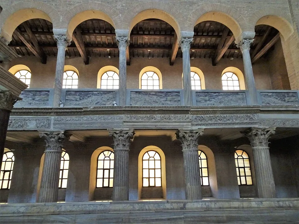 Two tiers of marble columns with carved capitals, the lower ones carrying a richly carved horizontal beam and the upper ones carrying round arches under a timber roof, with arched windows in the wall behind.
