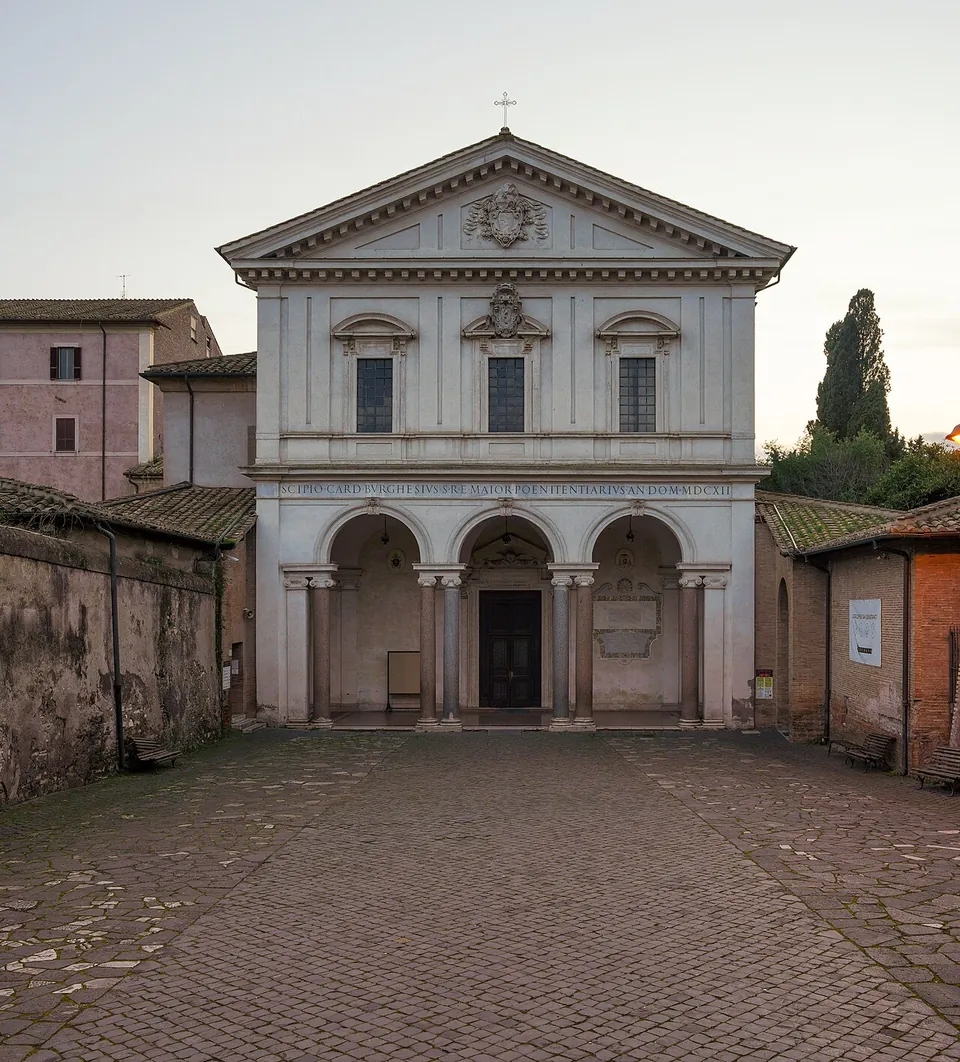 A white church front of two storeys with a triangular pediment, three arches on columns forming a portico below and a Latin inscription across the frieze, facing a cobbled forecourt between old walls.