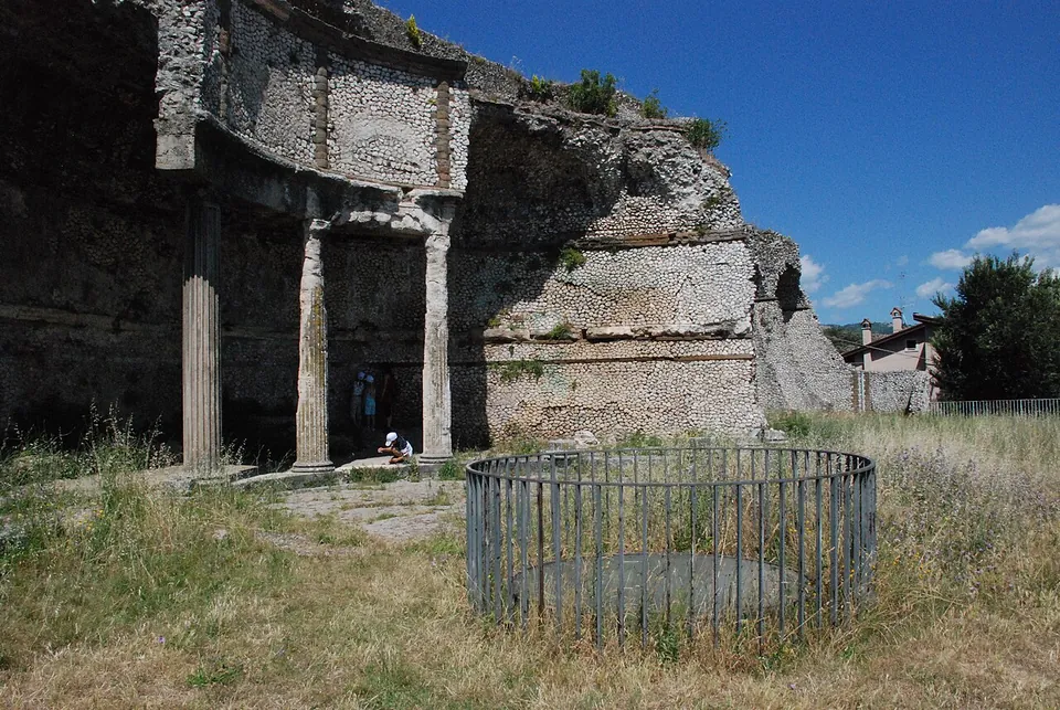 A curved recess cut into a massive wall of small stones, its front carried on columns under a curved stone beam, with two people sitting inside, dry grass in front and a round railing around an opening in the ground.