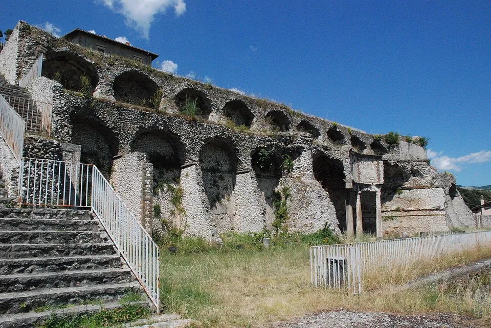 A wide stone stairway with metal railings climbing between two terrace walls of small rough stones, each pierced by tall arched niches, towards a pale building at the top against a blue sky.