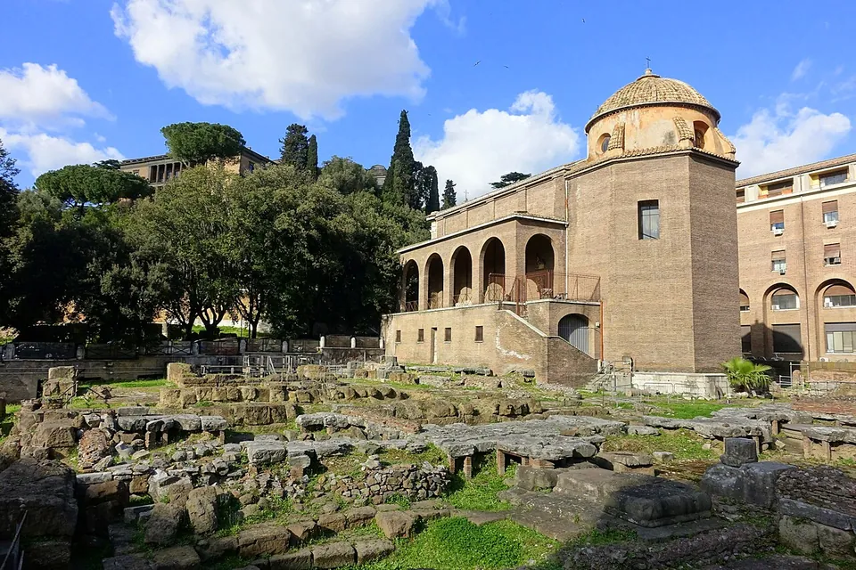 Low excavated walls and scattered blocks of grey stone across a sunken, grassy enclosure, with a small brick church with a domed lantern and an arcaded porch behind them under a blue sky.