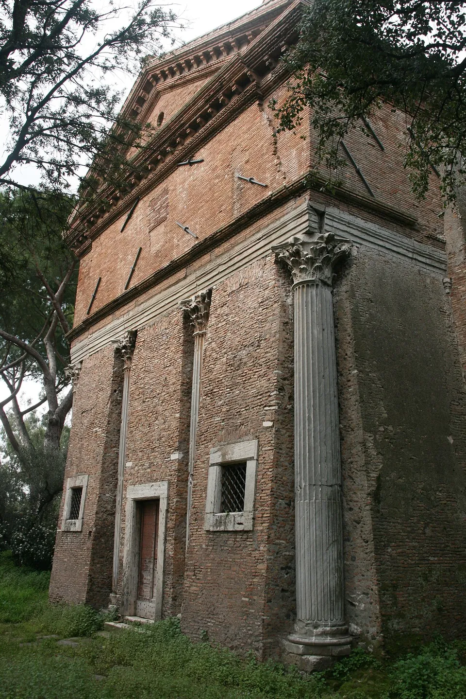 A tall brick building with a triangular pediment, fluted marble columns with ornate capitals built into its brick walls, two small barred windows and a narrow door, under overhanging trees.