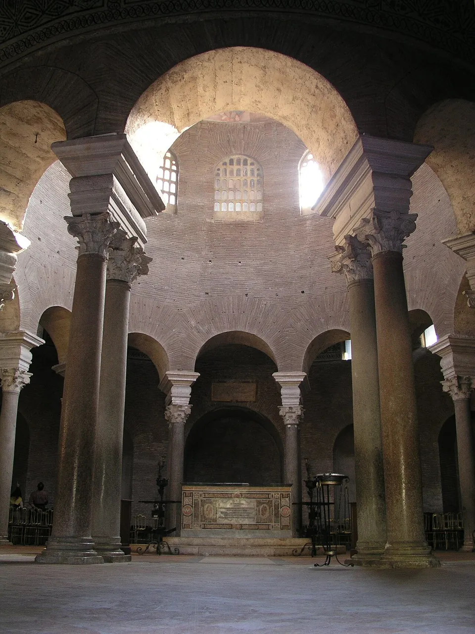 Tall paired columns with ornate capitals carrying round arches around a central space, daylight falling from arched windows in the bare brick drum above, and a low altar before a niche on the far side.
