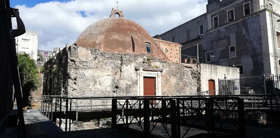 A square building of rough grey stone with a framed doorway, crowned by a broad reddish dome, seen across a railed metal walkway laid over excavated ground, with tall city buildings behind.