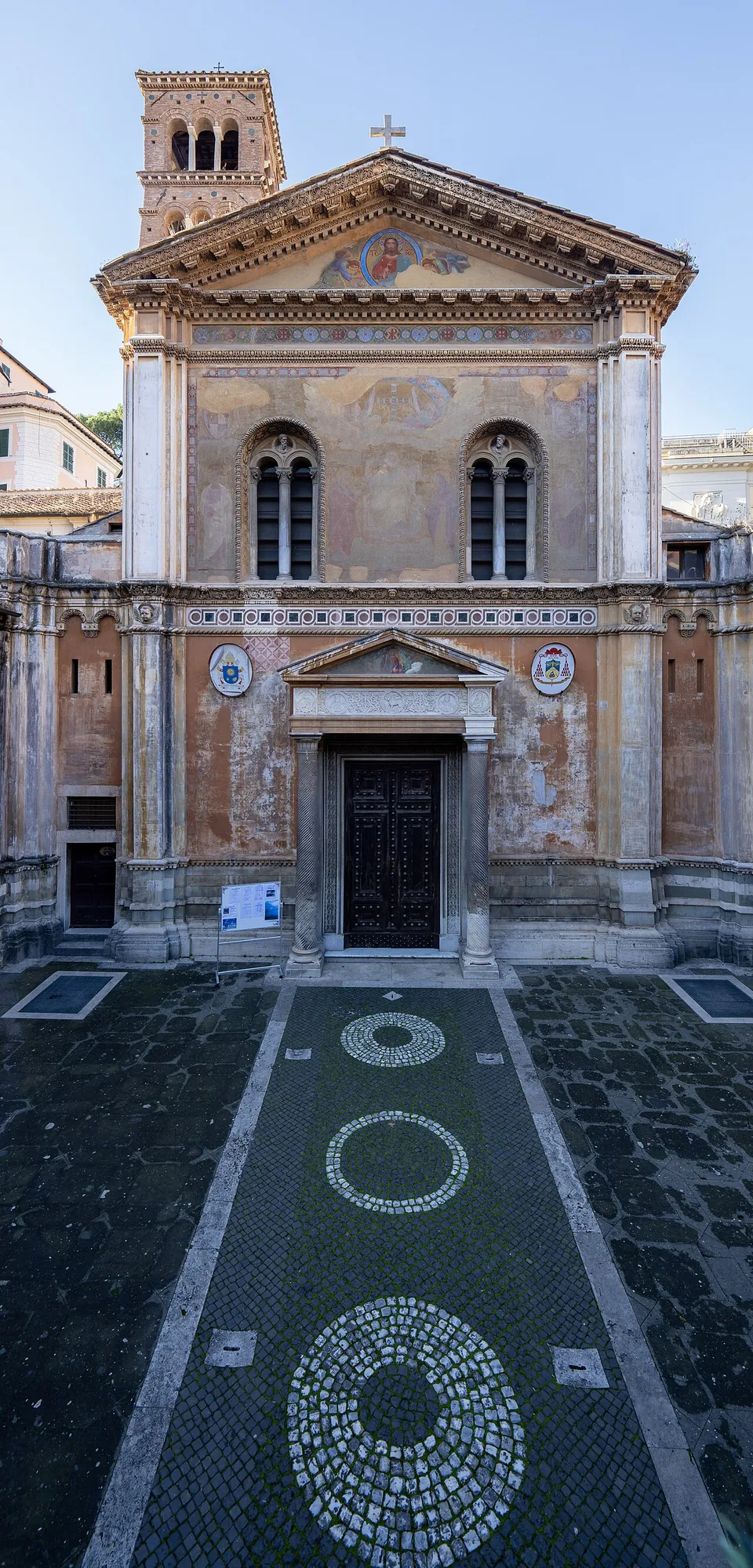 A church façade seen from a paved courtyard laid with circular patterns in white stone, with a pedimented doorway, two arched windows, painted roundels and a brick bell tower rising behind the gable.