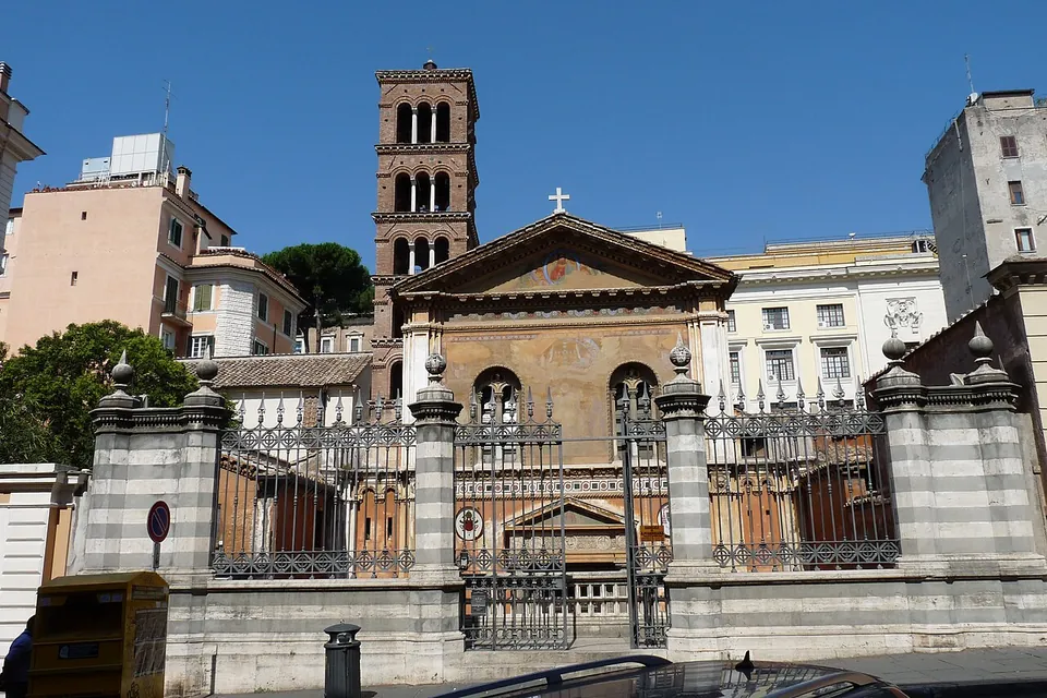 A street-side wall of grey-and-white banded stone with iron railings and a gate, through which the pedimented façade of a church can be seen standing lower than the street, with a tall brick bell tower behind it.