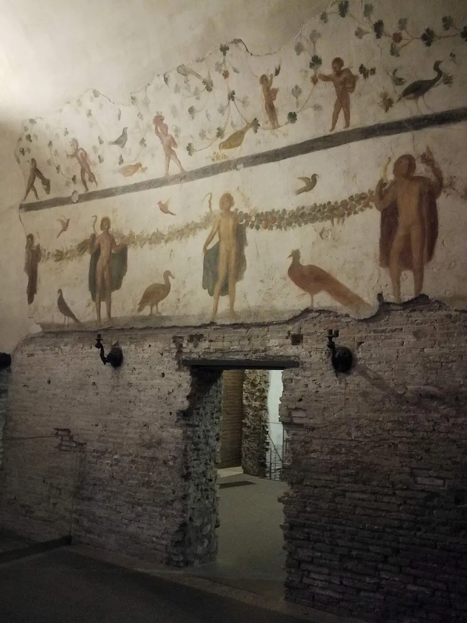 A room whose upper walls are painted on a pale ground with standing youthful figures linked by garlands, birds including a peacock, and a frieze of small figures among vine leaves, above lower walls of bare brick with a doorway.