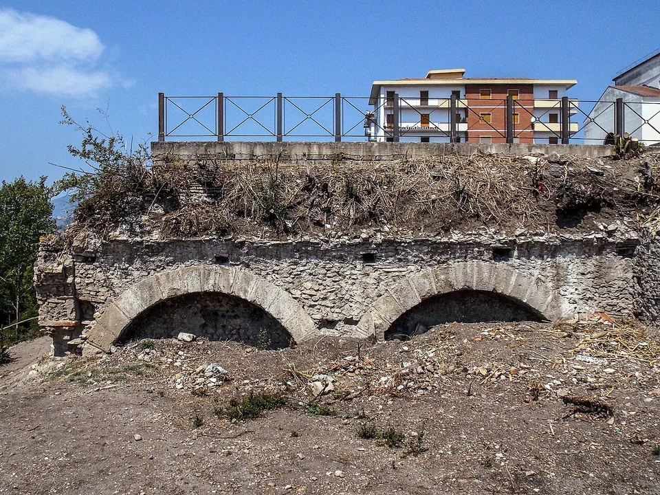 Two low brick arches in a stone wall, their openings half filled with earth, under a bank of dry grass topped by a railing, with a block of flats behind.