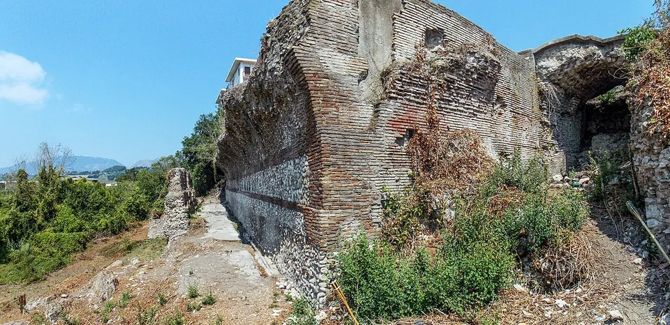 A tall wall faced with courses of red brick, rising from weeds and rubble, with a dark vaulted opening at its right-hand end and a strip of hills and trees on the left.