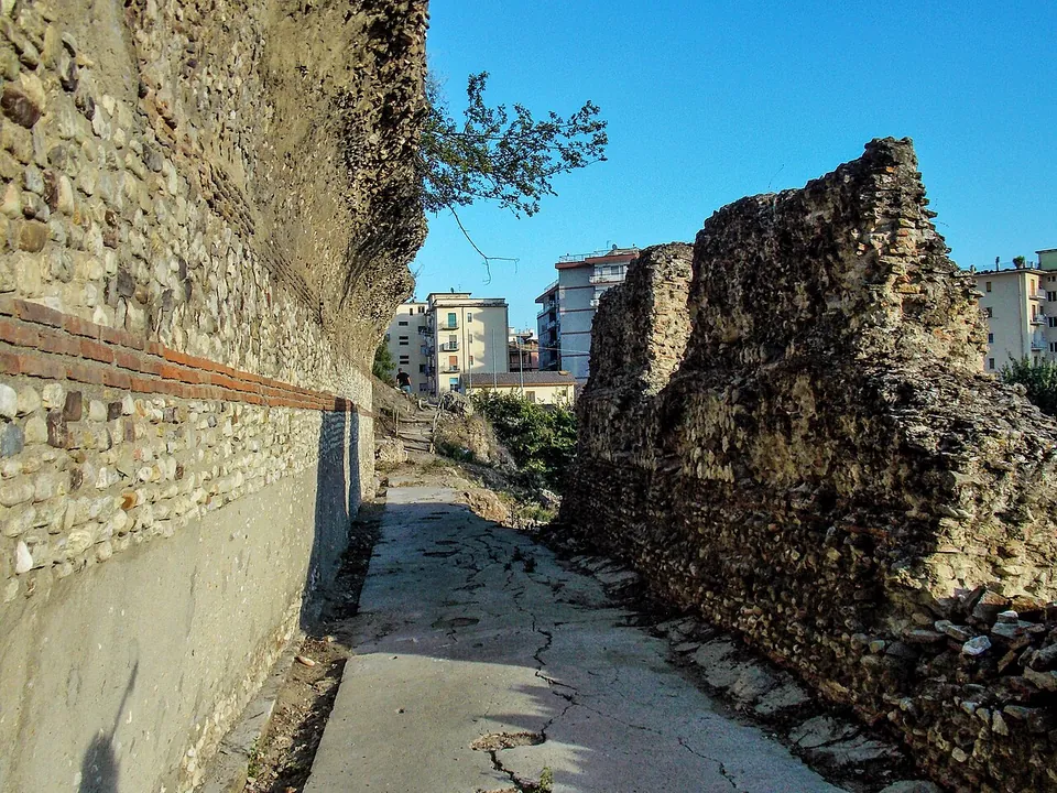 A concrete path running between a rough rubble wall cut into a bank on the left, with a band of brick near its foot, and a long ruined wall of stone and brick on the right, with blocks of flats behind.