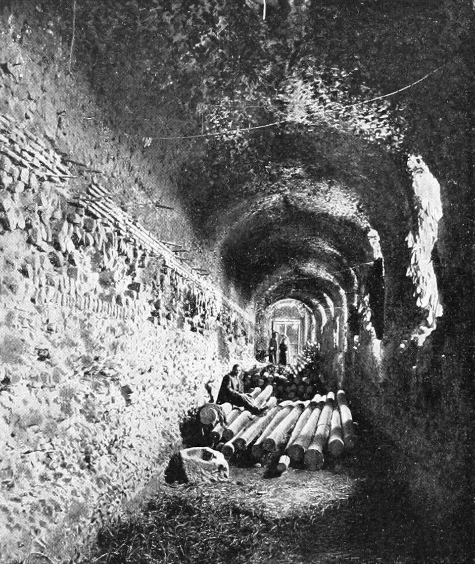 A black-and-white photograph down a long, dark barrel-vaulted gallery, its rough walls lit from openings on the left, with wooden beams stacked on the floor and a figure sitting among them.