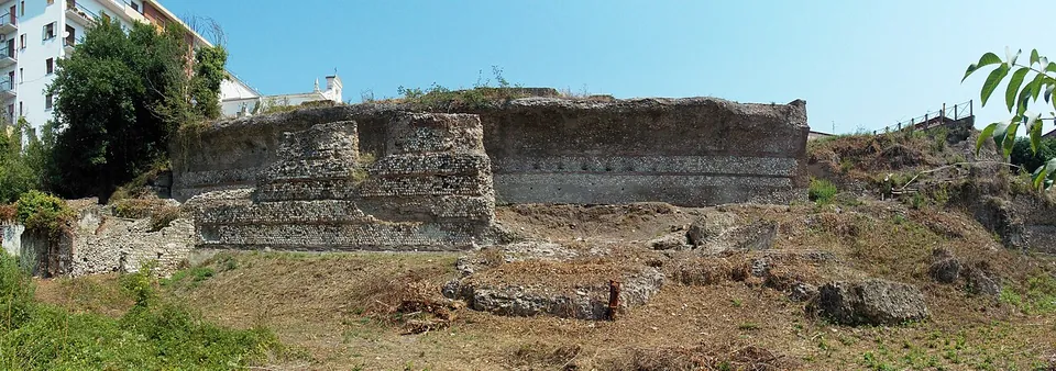 A wide view across rough grass to a long, low ruined wall of stone and brick standing along the foot of a slope, with blocks of fallen masonry in front of it and modern buildings at the left edge.