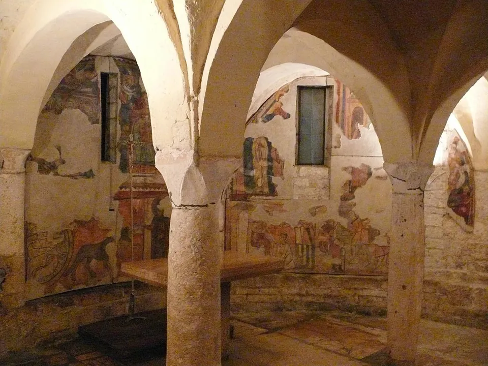 A low vaulted crypt with stone columns and arches, its walls and vaults covered with patches of faded painting.