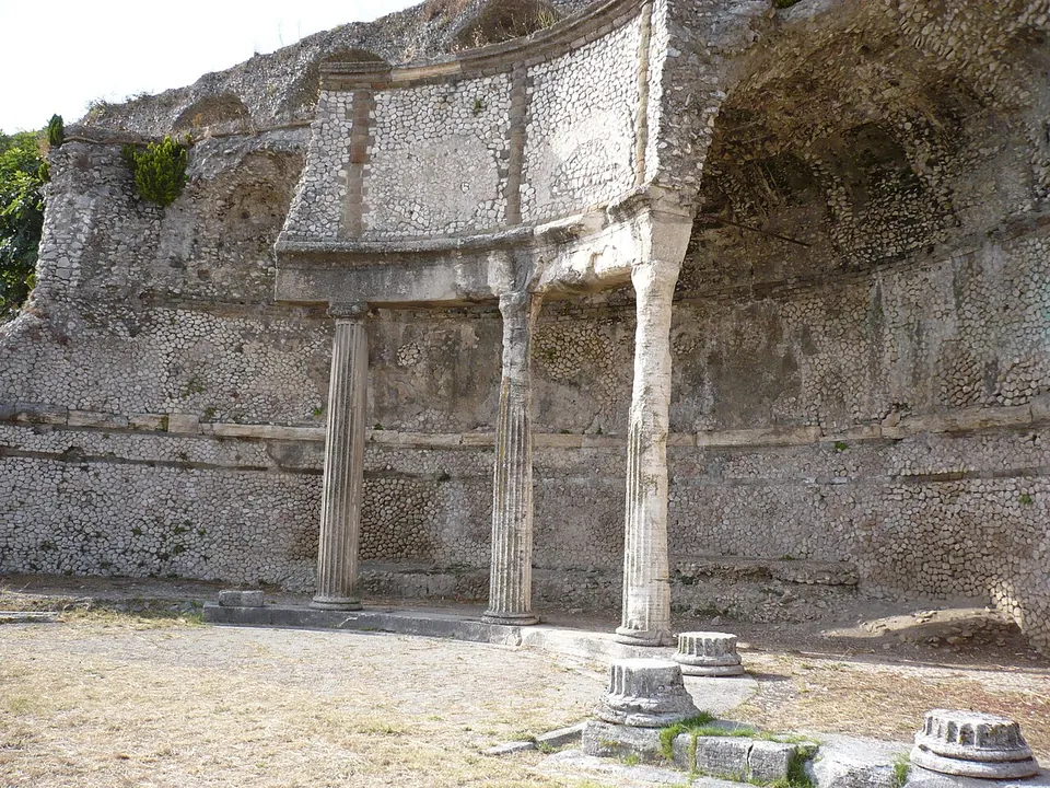 A curved wall of small rough stones with a moulded band along it, fronted by three slender columns that carry a curved stone beam and a panelled upper wall, with column bases and fragments lying on the gravel in front.