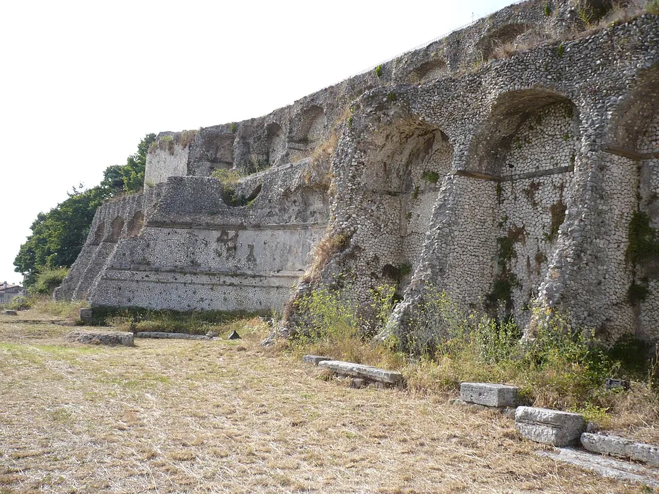 A long, high terrace wall of small rough stones broken into a row of tall arched niches with heavy piers between them, beside a dry, grassy terrace scattered with stone blocks.