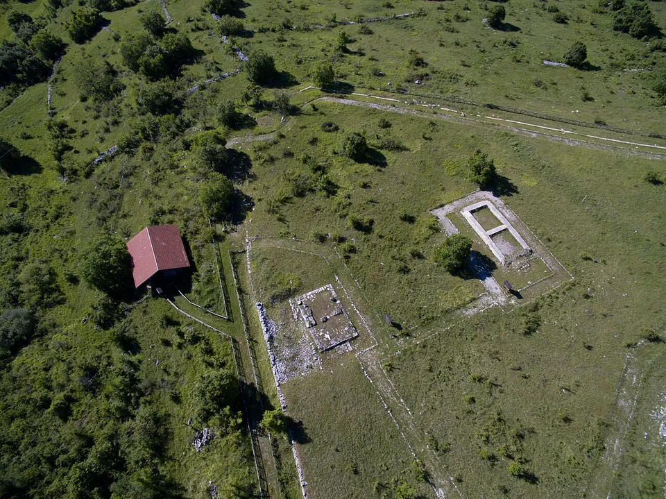 An aerial view straight down on a green upland plateau with scattered trees, where pale lines of low stone walls mark out rectangular enclosures and the plans of buildings, with a red-roofed shelter beside a path on the left.