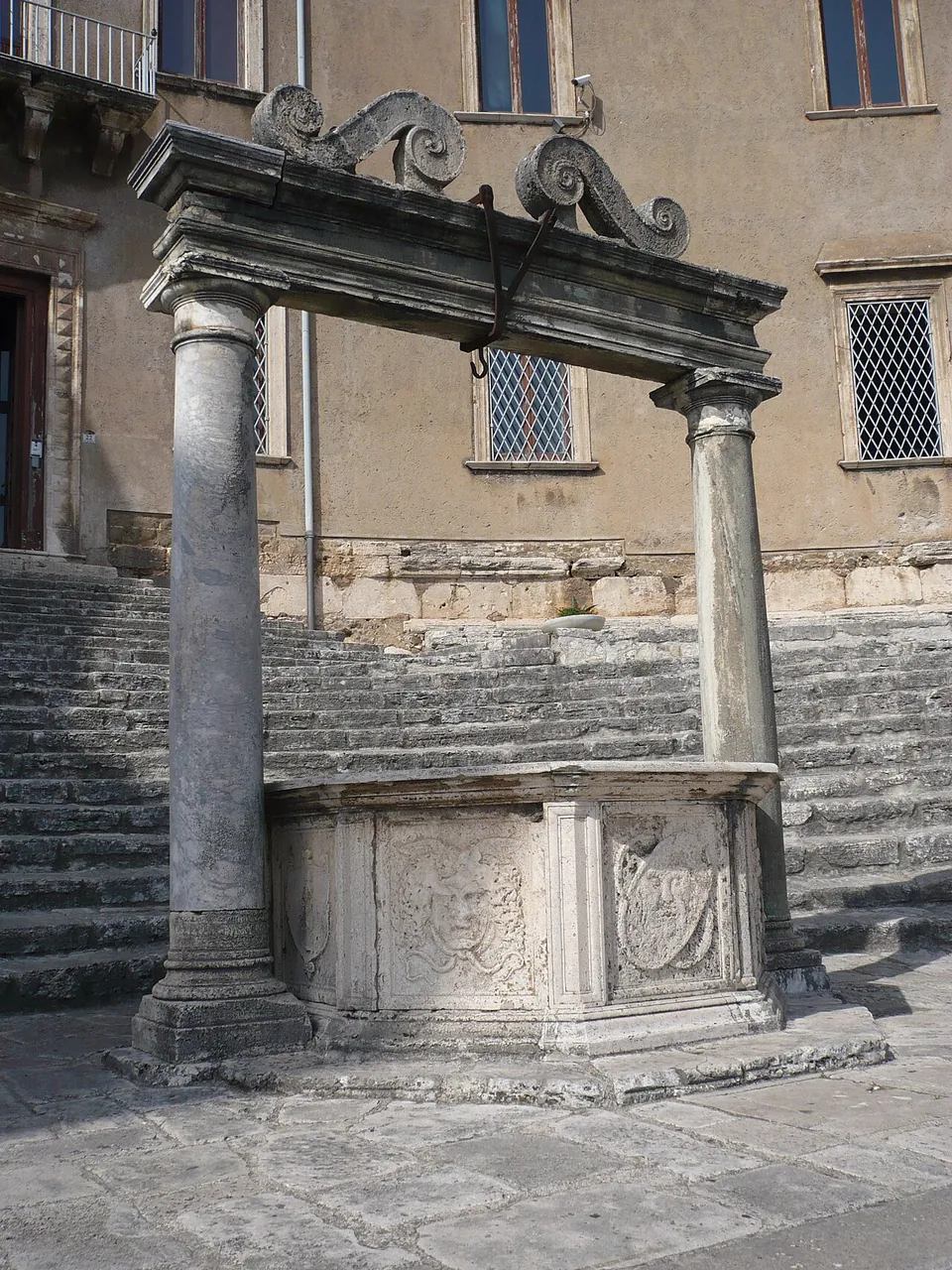 A carved stone well-head between two columns that carry a lintel topped with stone scrolls, standing on paving in front of a wide flight of stone steps that leads up to the plain facade of a palace.