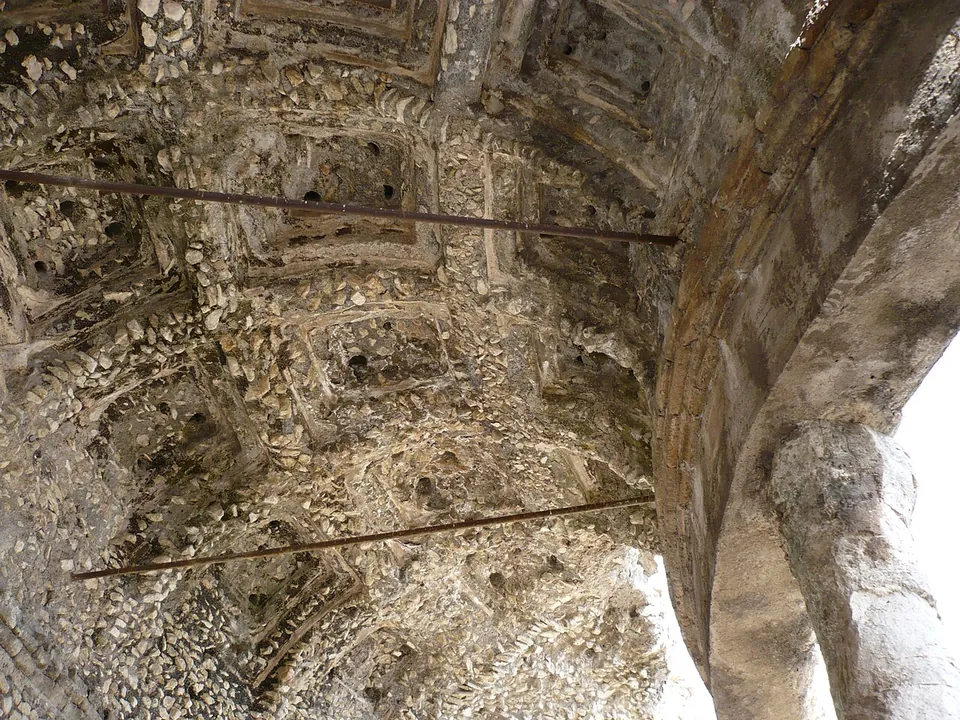 Looking up into a curved vault patterned with deep square coffers, built of small stones set in mortar, with two iron tie-rods across it and the edge of an arch on the right.