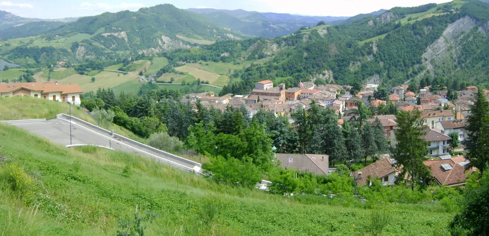 The town seen from a grassy slope above it, a cluster of red-tiled roofs among trees in a green valley ringed by wooded and farmed hills.