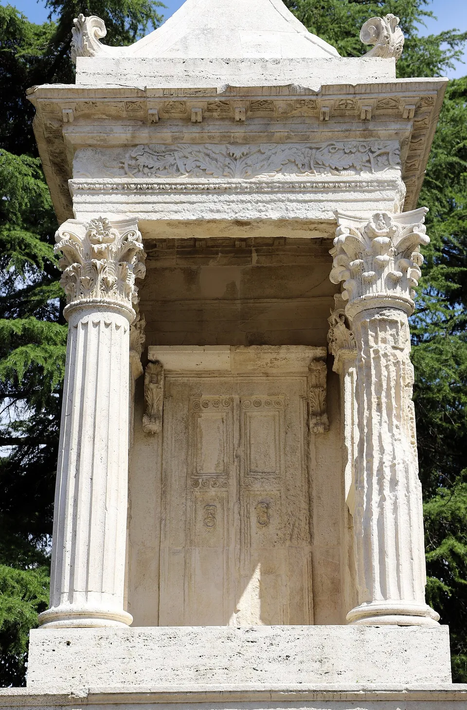 Close view of the open upper storey, where two fluted columns with leafy capitals frame a stone panel carved as a double door, under a cornice with a carved frieze.