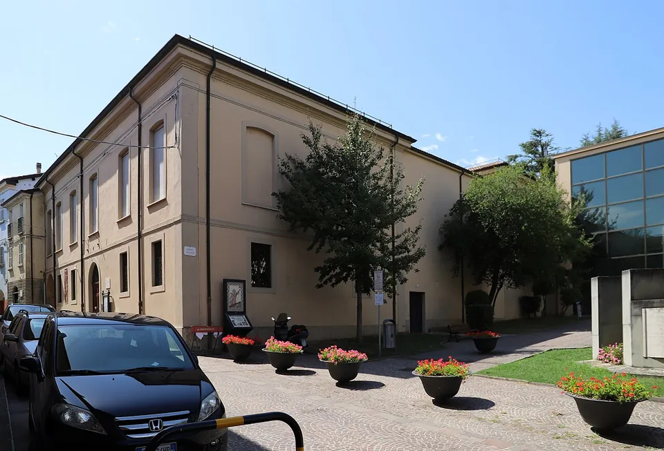 The plain two-storey front of the museum along a cobbled street, with flower pots, a stone column and a block on a plinth in a small lawn, and a tall glazed hall beside it.