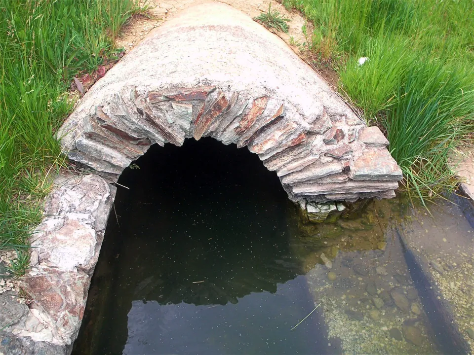 A low barrel vault of thin stones laid on edge over a channel of dark water, set into a grassy bank.