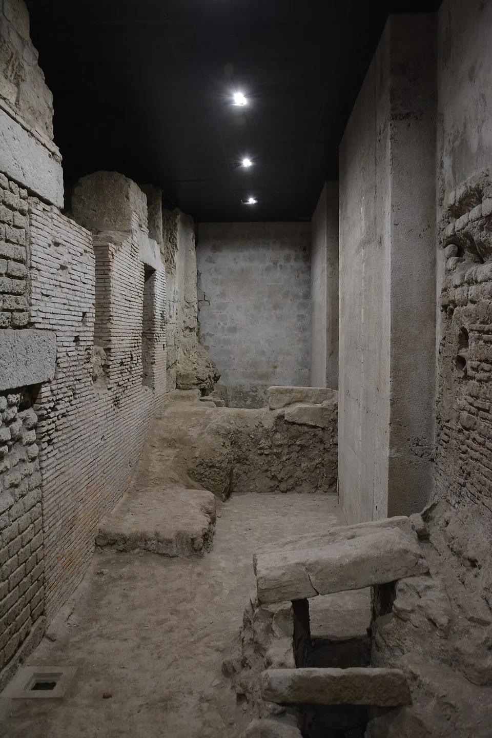 A narrow excavated space between walls of brick and squared tuff blocks, with low walls and flat stone slabs set on the floor, lit from a black ceiling.