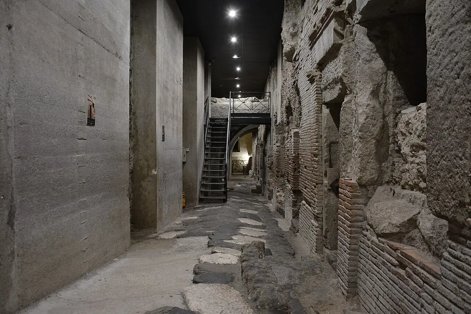 An excavated street paved with irregular dark stone blocks runs between ancient walls of brick and tuff, with doorways opening off it on the right and a smooth modern wall on the left, lit by a row of ceiling lamps, with a steel staircase at the far end.