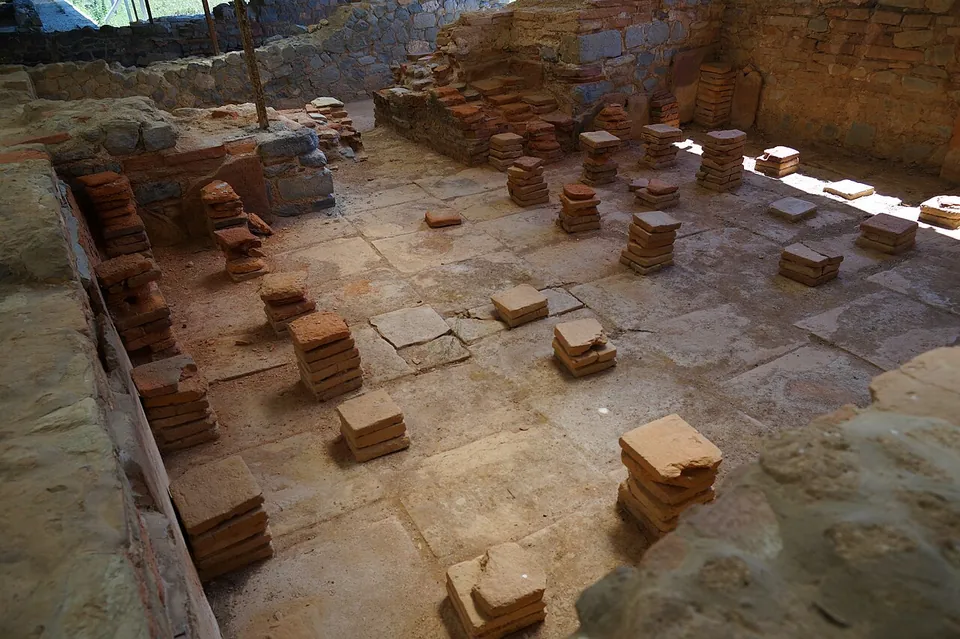 A sunken room with a floor of large worn tiles, scattered with short stacks of square red bricks, enclosed by low walls of stone and brick.