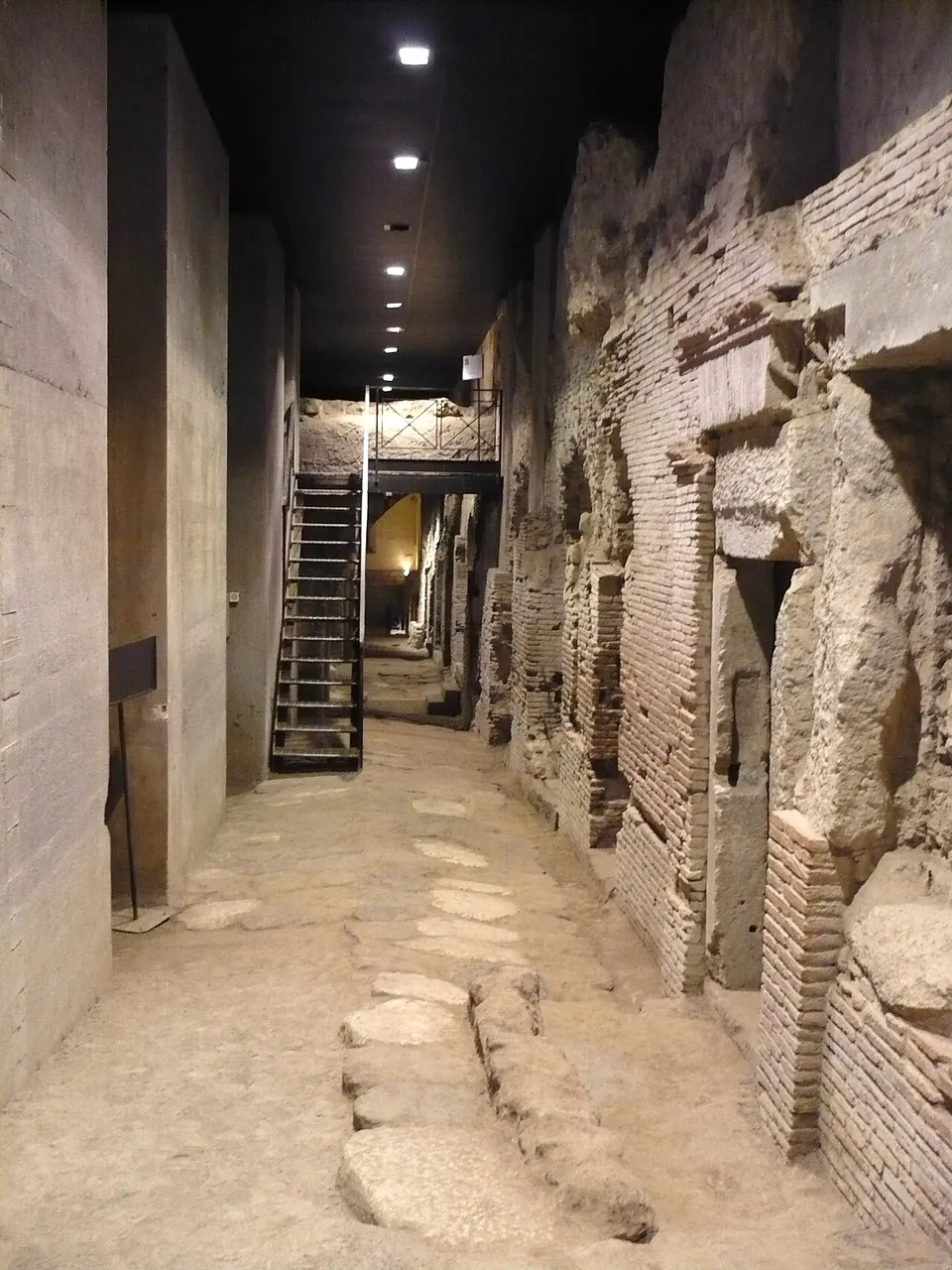 An excavated street paved with large irregular stone blocks runs between a smooth modern concrete wall on the left and ancient walls of brick and tuff on the right, with doorways opening off them, under a row of ceiling lamps; a steel staircase climbs at the far end.