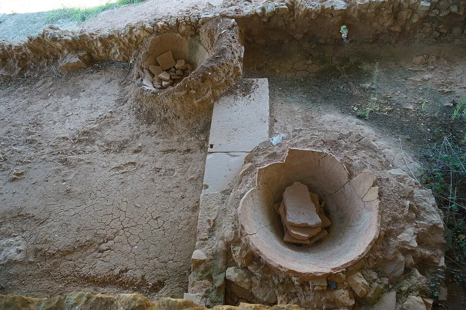 Two large round terracotta jars set into the bare earth floor, their rims at ground level and broken pieces lying inside them, beside a stone slab.